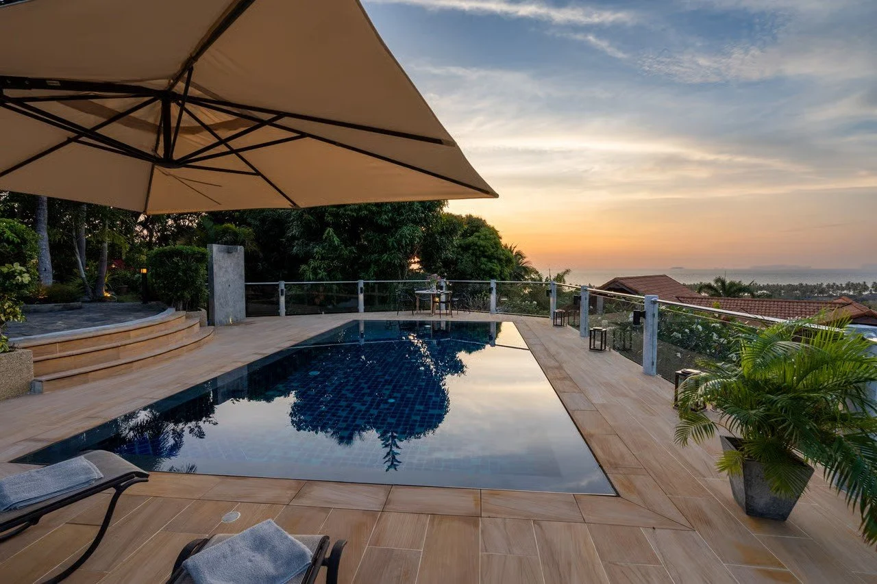 A luxurious poolside area during sunset with a rectangular pool, a large beige umbrella, lounge chairs with towels, potted plants, and a railing overlooking trees, rooftops, and the ocean in the distance.