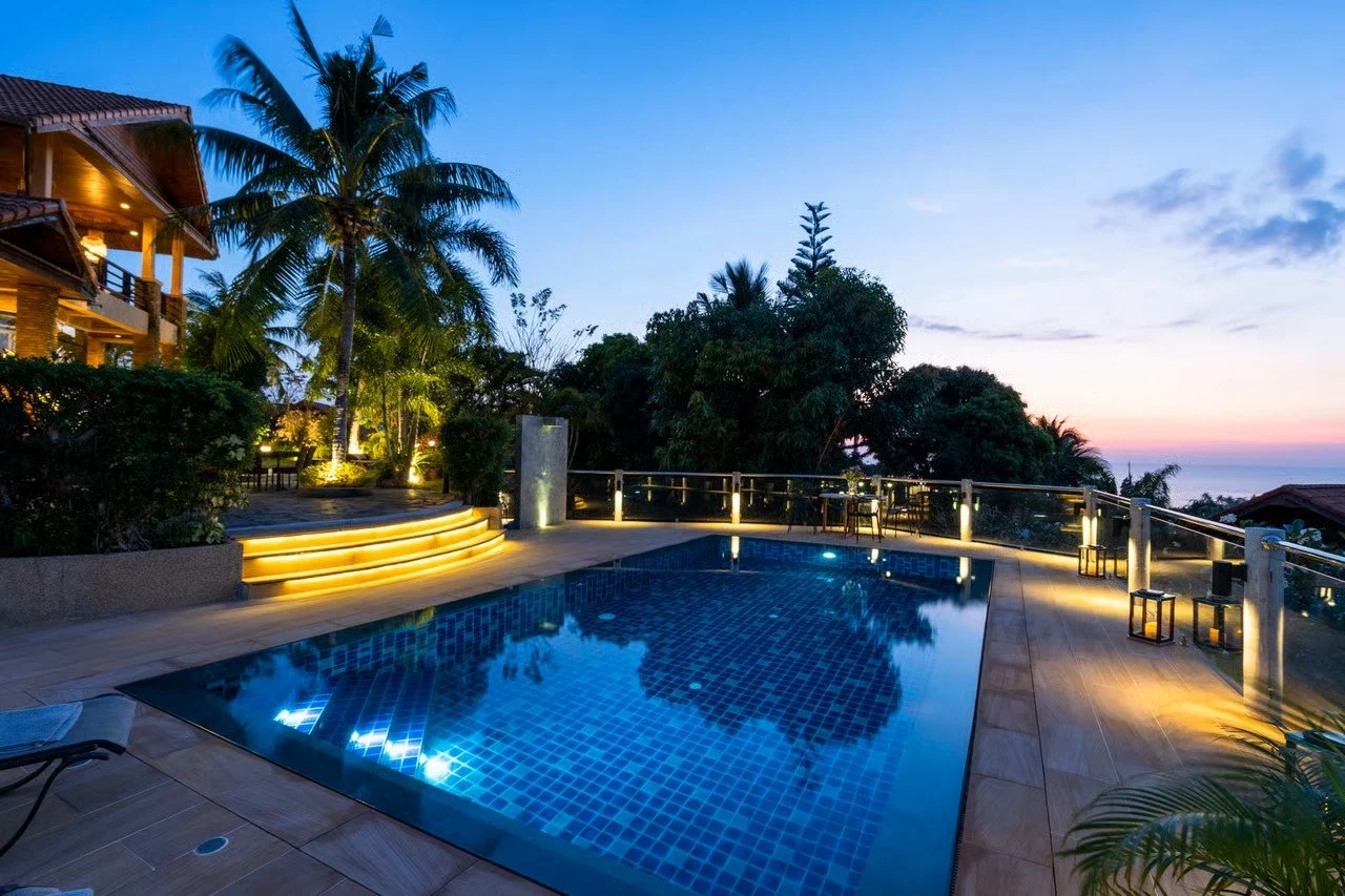An outdoor swimming pool with underwater lights, surrounded by a deck with lanterns and a glass railing, overlooking a lush garden and a sunset over the ocean.