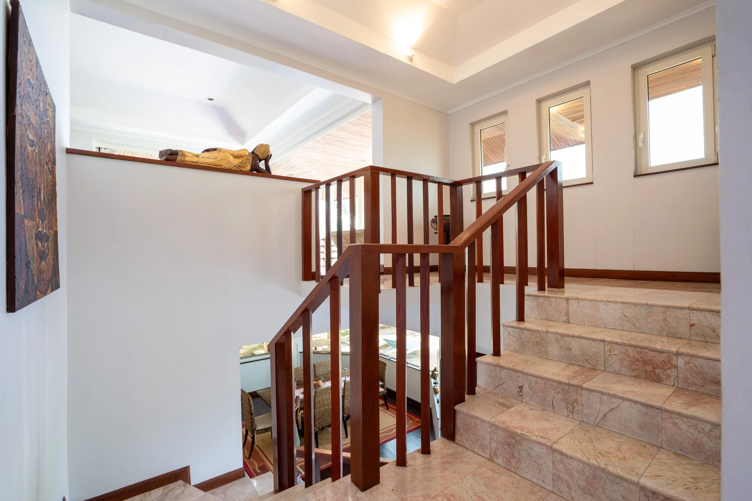 Interior view of a staircase with marble steps, wooden railing, and large windows, leading to an upper level with white walls and ceiling.
