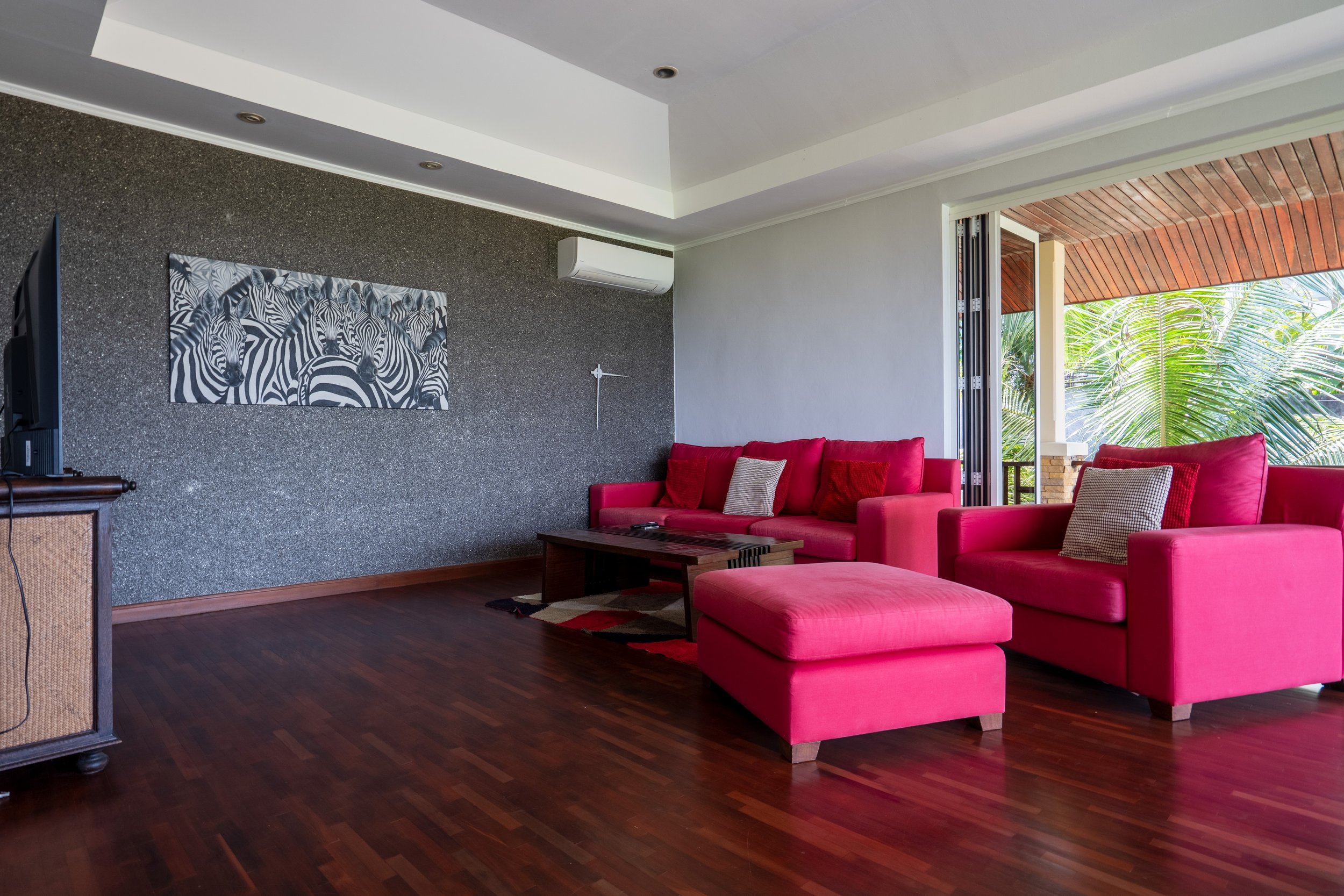 Living room with pink sofas, a dark wooden coffee table, a black and white zebra artwork on a textured wall, and large sliding glass doors opening to a balcony with greenery.