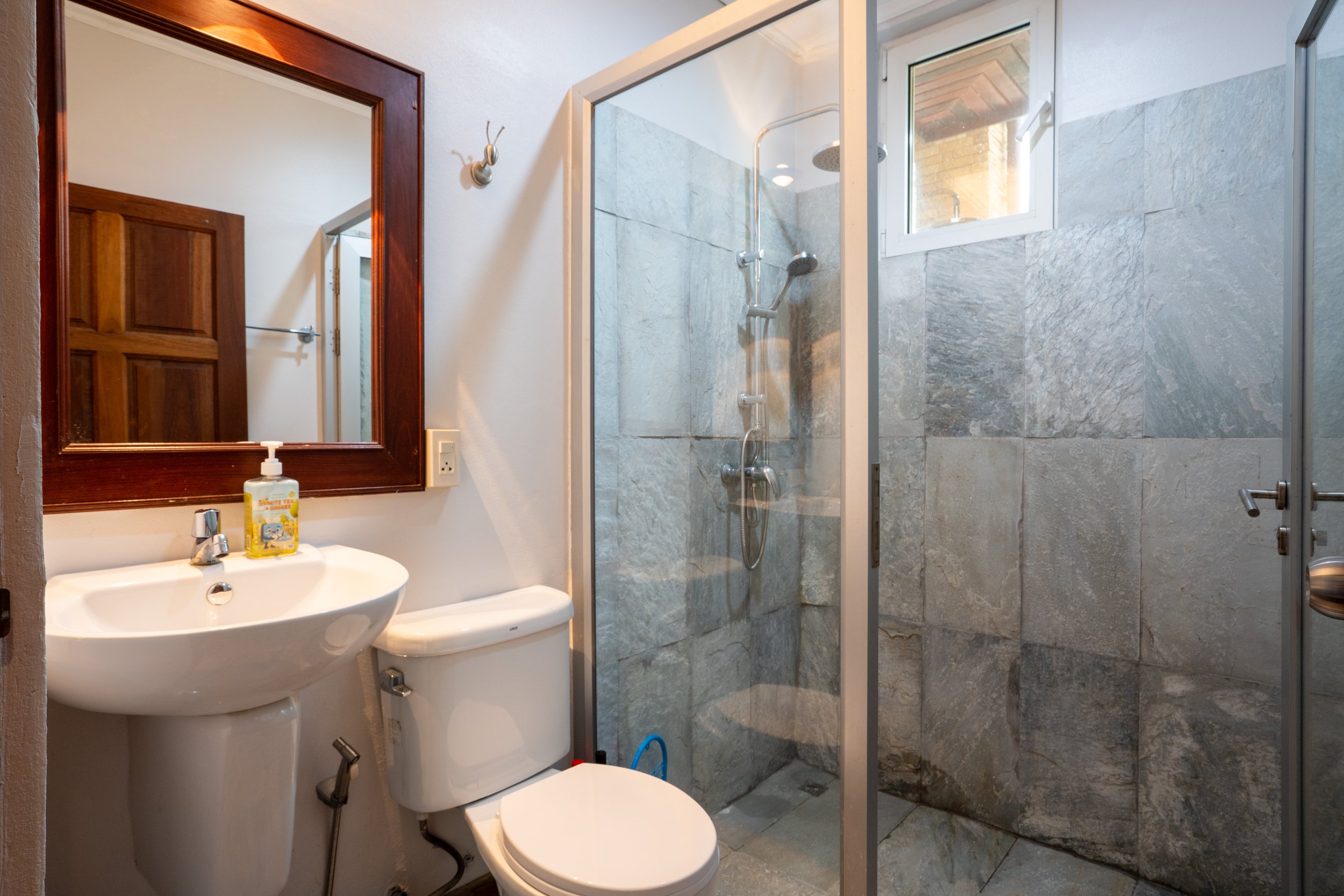 Bathroom with a sink, mirror, toilet, and a glass-enclosed shower with a small window, wood door, and tiled walls.