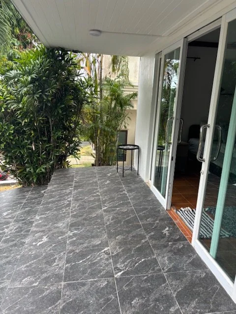 A covered outdoor patio with gray tiled flooring, a small black stool, and sliding glass doors leading inside a house, with greenery visible in the background.