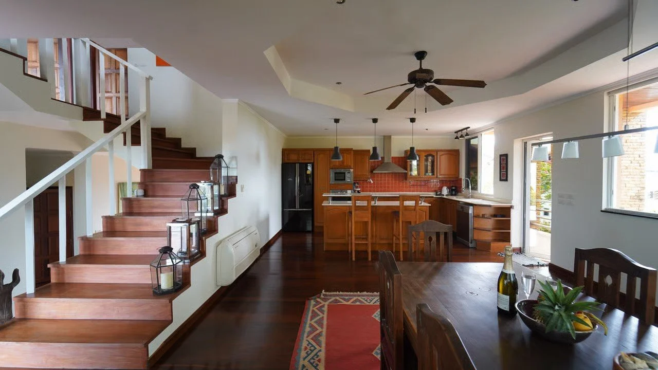 Open-concept living space with a staircase, kitchen, dining table with a bottle of champagne and glasses, and natural light from large windows.