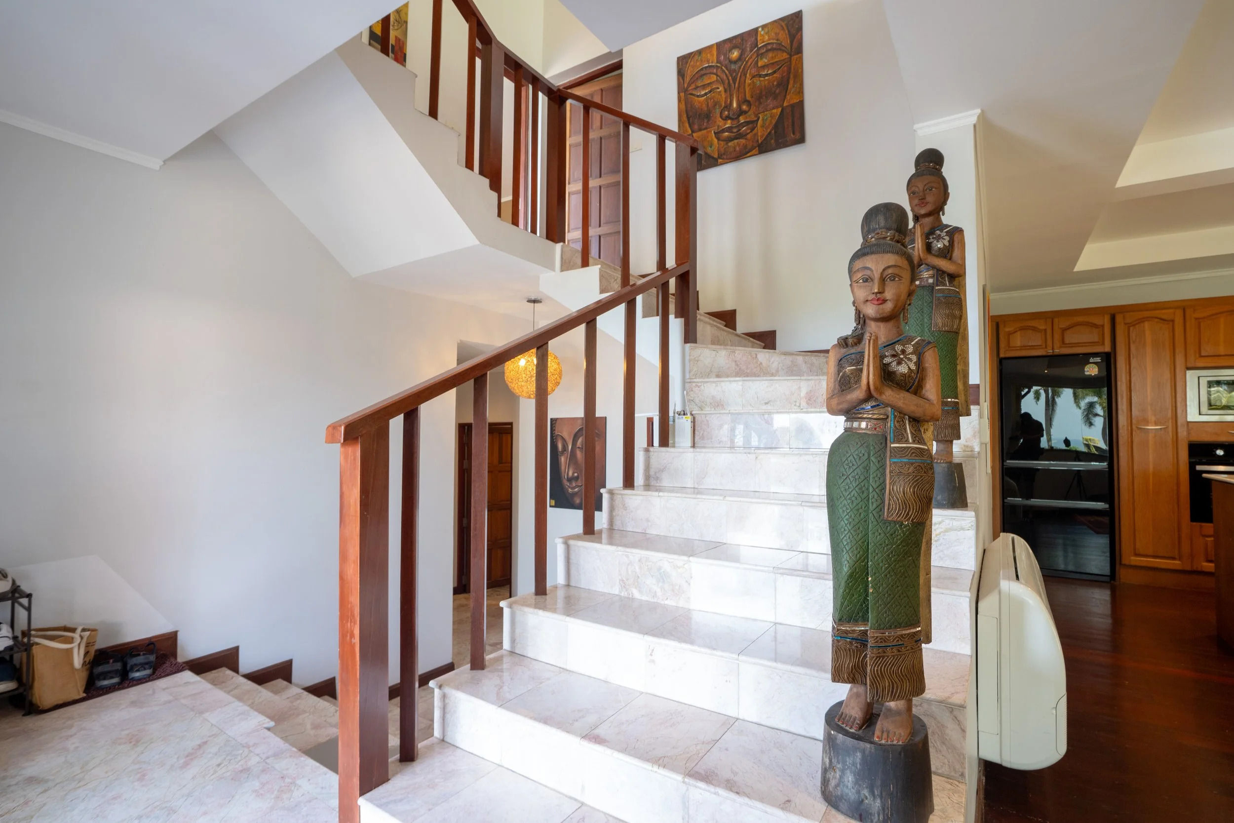 Decorative wooden statues of women with hands in prayer pose near the staircase in a home interior. Artwork of a serene face hangs on the wall above the stairs.