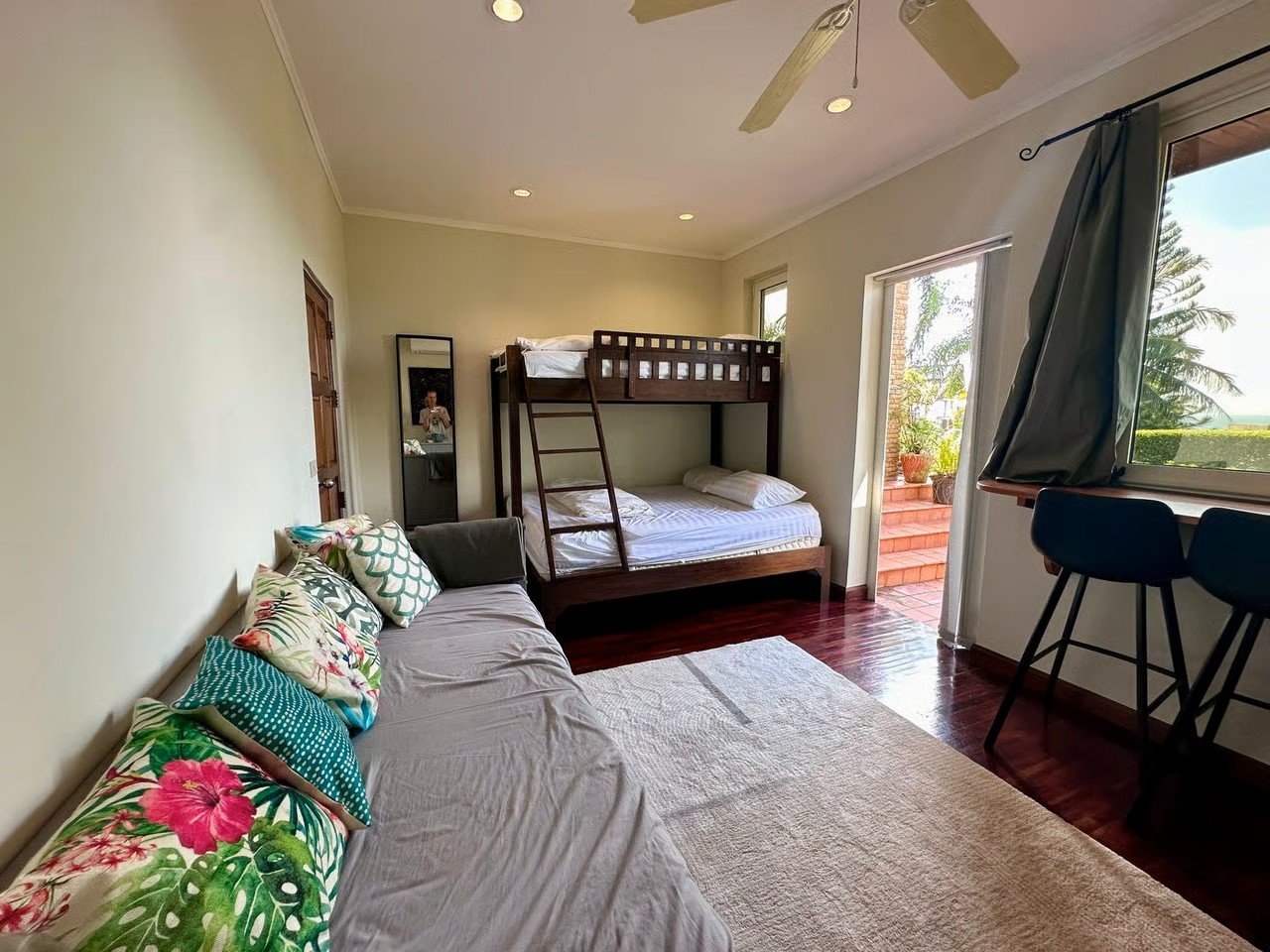 A cozy room with a large sofa decorated with colorful tropical print pillows along one wall, a bunk bed with a ladder next to a small mirror, and a window and door leading to outdoor steps with potted plants. The room has wooden flooring, a ceiling f
