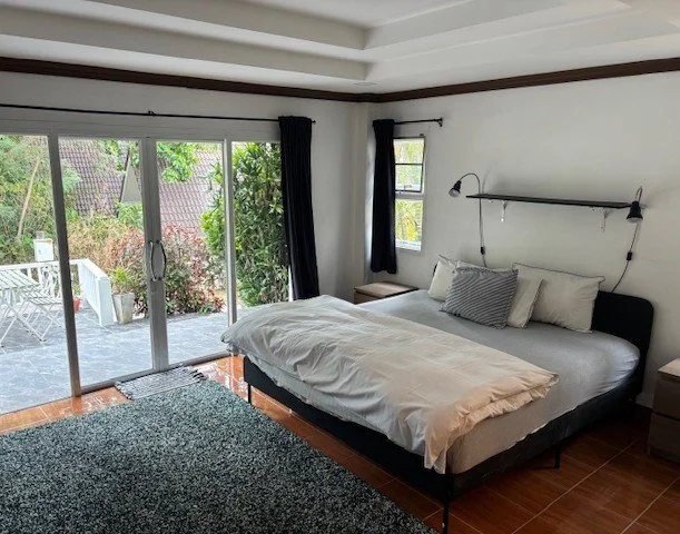 Bedroom with large bed, dark headboard, white bedding, gray pillow, two nightstands, black curtains, sliding glass door leading to patio with outdoor furniture, small window with curtain, and a textured rug on wooden floor.