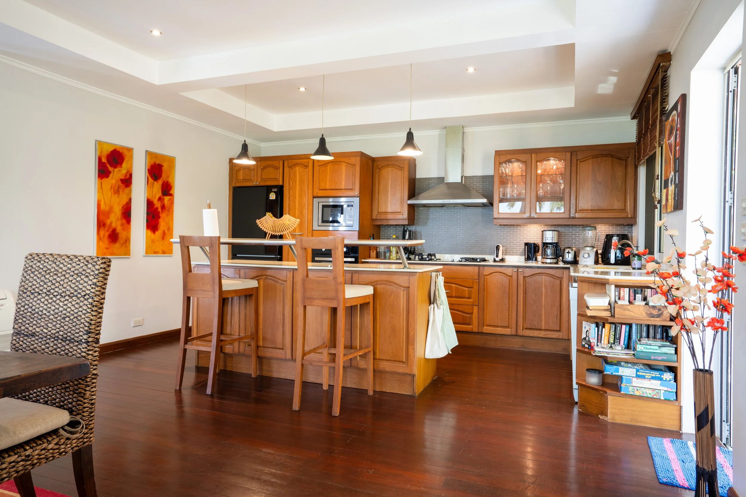 A kitchen with wooden cabinets, a kitchen island with high chairs, a black refrigerator, stainless steel stove and microwave, and various small appliances on the counters. There is a bookshelf with books and decorative items, and a window with wooden