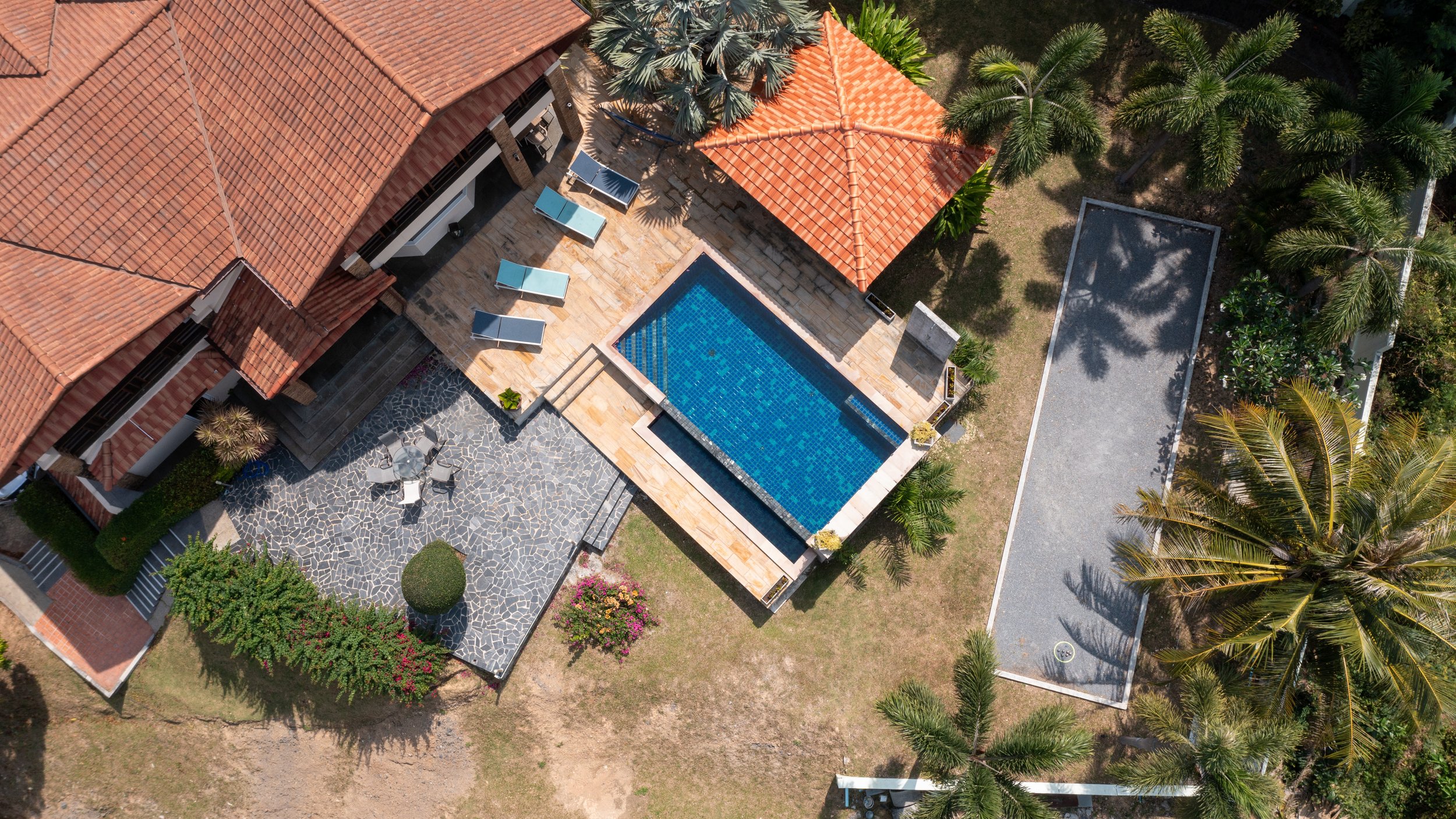 An aerial view of a backyard featuring a swimming pool with blue tile lining, a stone patio with lounge chairs, a small wooden gazebo with a tiled roof, and grassy areas with palm trees and plants.