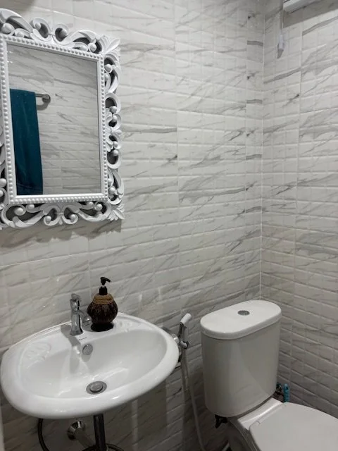 A small bathroom with a white sink, a decorative framed mirror, and a toilet. The walls are tiled with white marble-patterned tiles.
