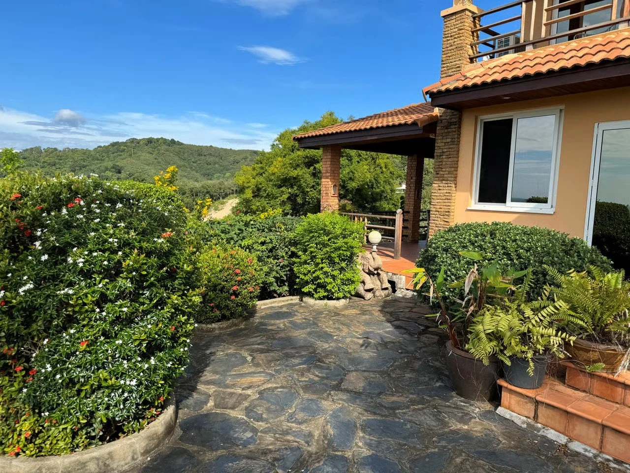 Stone patio with lush green bushes and potted plants outside a house built with brick and stucco, surrounded by trees and hills under a blue sky.