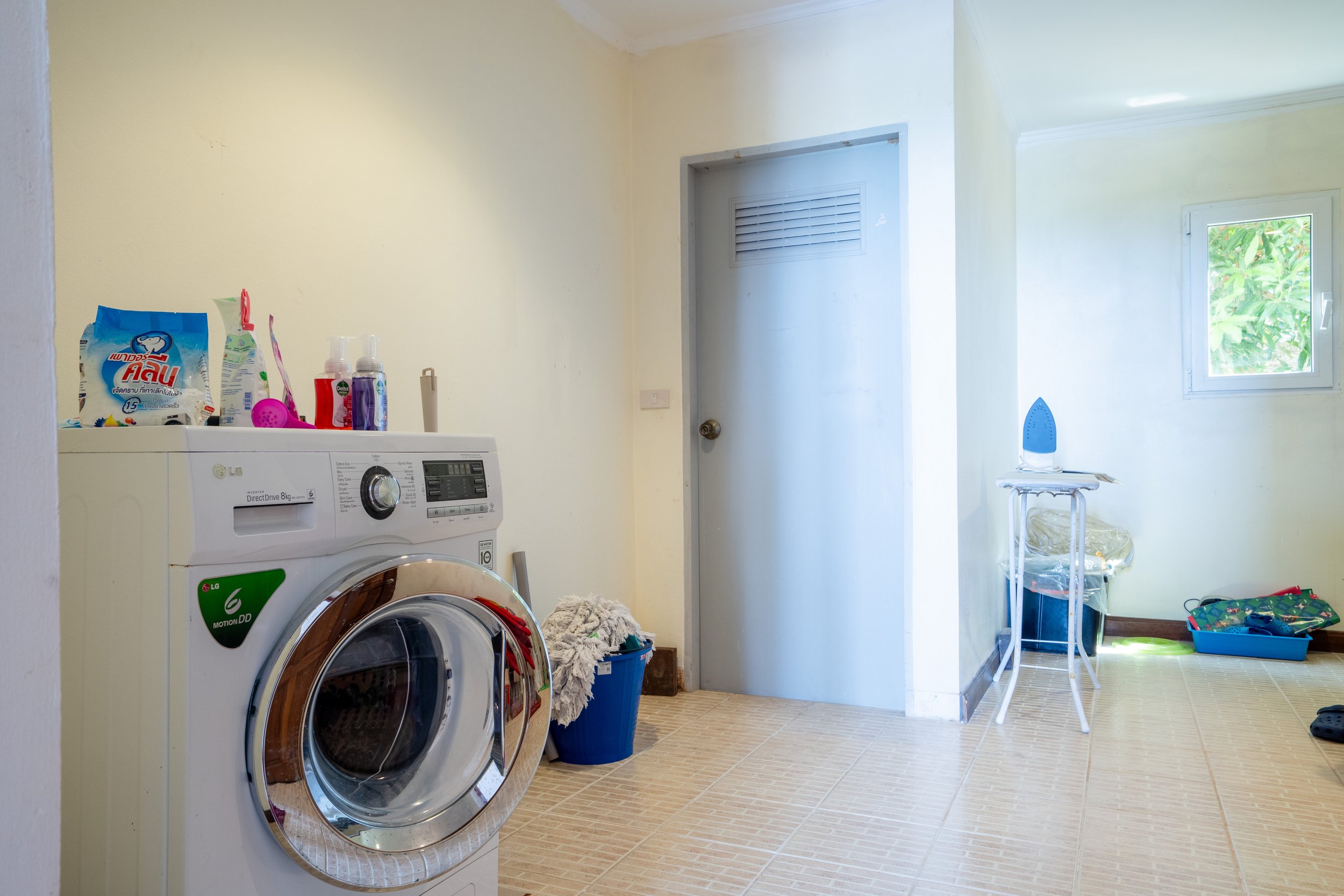 A laundry room with a washing machine, laundry supplies on top, a blue laundry basket, an iron on an ironing table, and a small window with greenery outside.