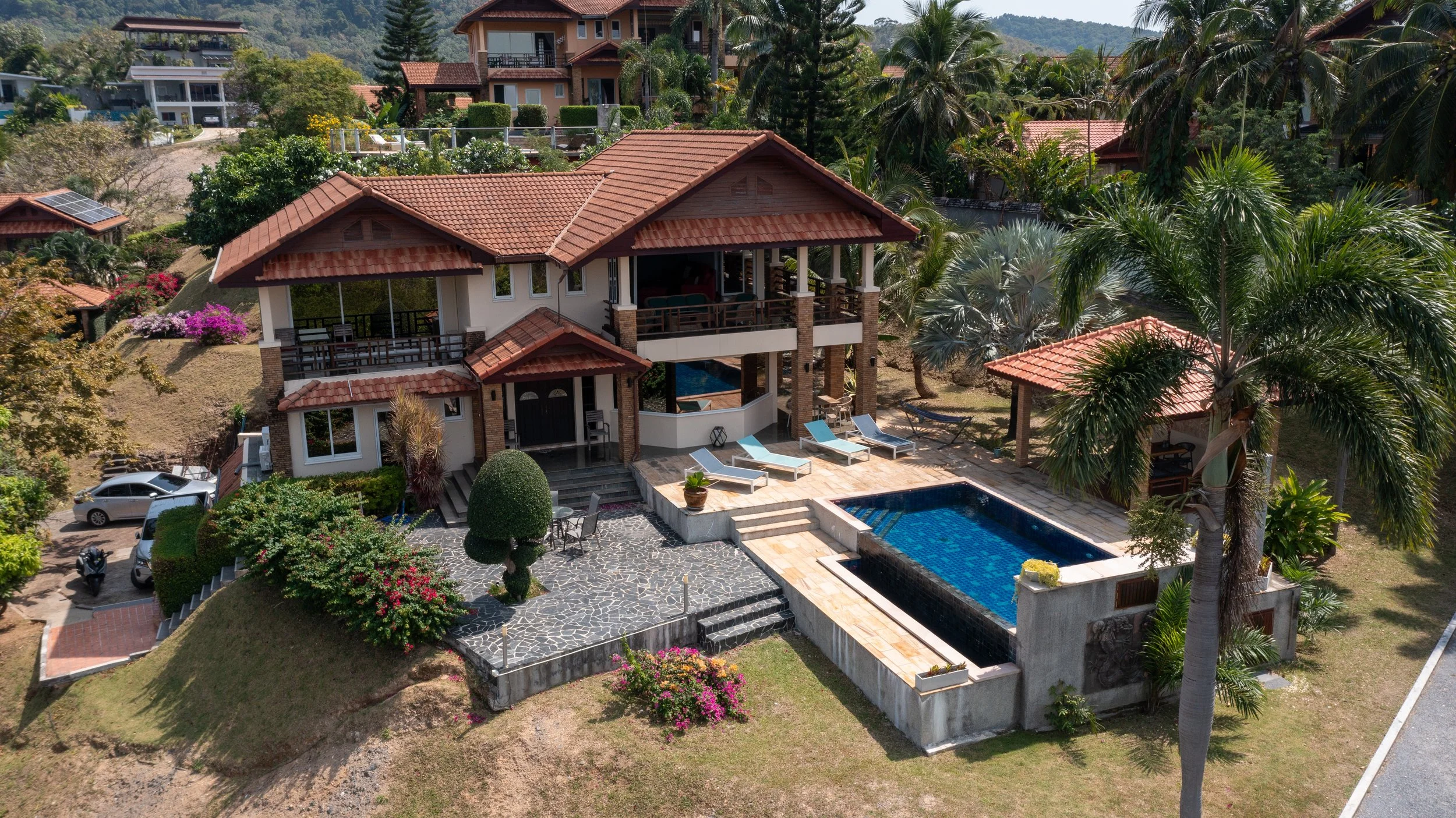 A large house with a red tiled roof, multiple balconies, and a swimming pool surrounded by a tiled patio with lounge chairs. The yard features well-maintained greenery, including palm trees, colorful flowering bushes, and a paved stone area with a ta