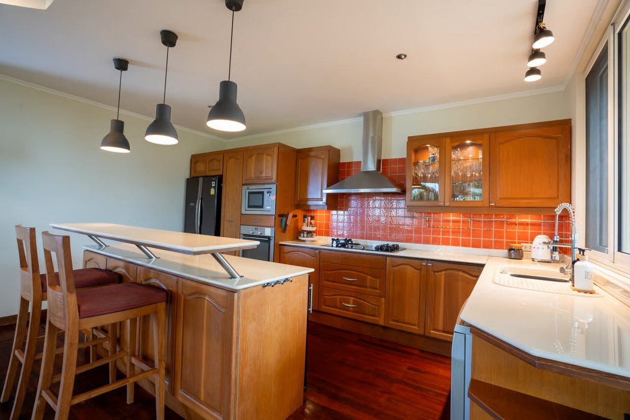 Kitchen with wooden cabinets, red tile backsplash, stainless steel appliances, and a white countertop. There is a small breakfast bar with three wooden chairs. Hanging pendant lights are above the breakfast bar, and there's a window with a view outsi