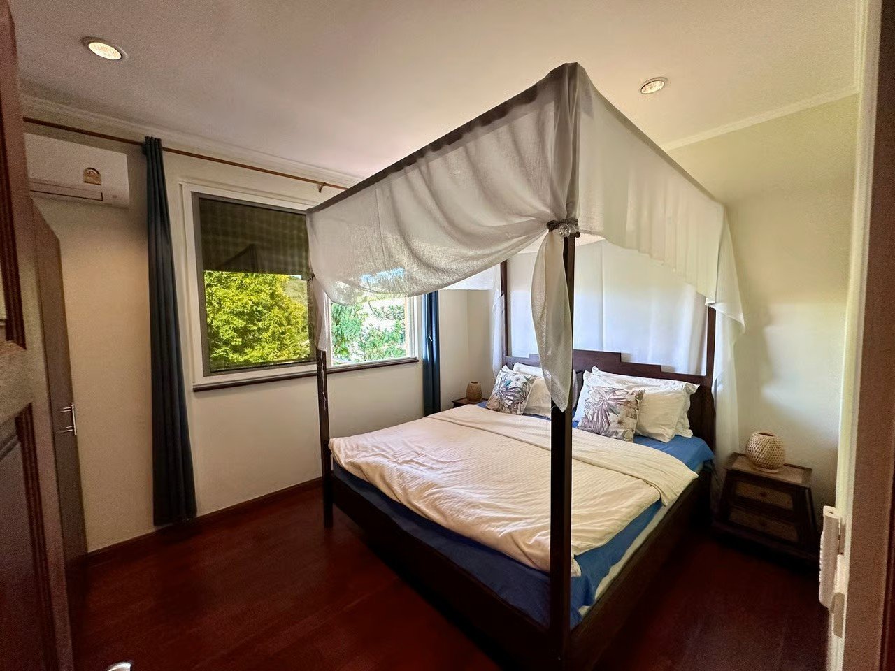 A bedroom with a four-poster bed and white canopy, two pillows, a side table with a basket, a window with view of green trees, and an air conditioning unit.