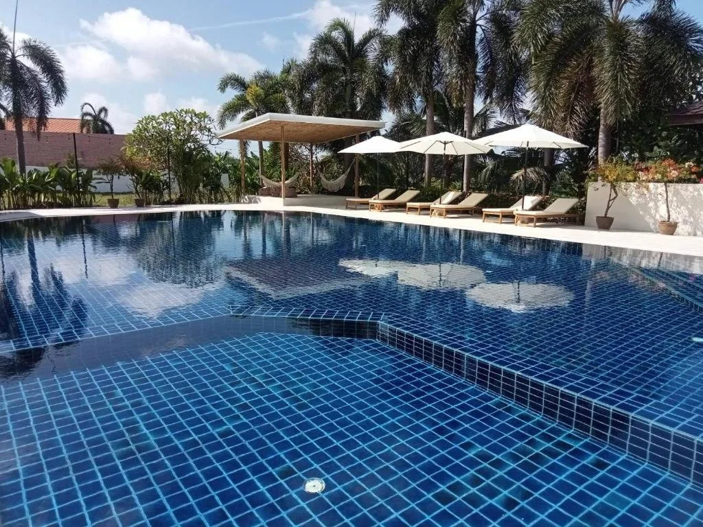 Outdoor swimming pool with blue tiles, surrounded by lounge chairs with umbrellas, palm trees, and a shaded cabana, under a partly cloudy sky.
