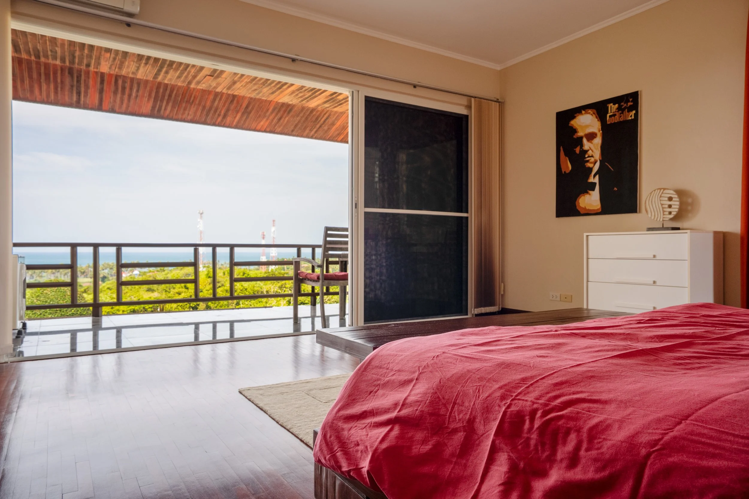 Bedroom with red bedspread, white dresser, wall art of 'The Godfather', sliding glass door open to a balcony with a view of trees and sky.