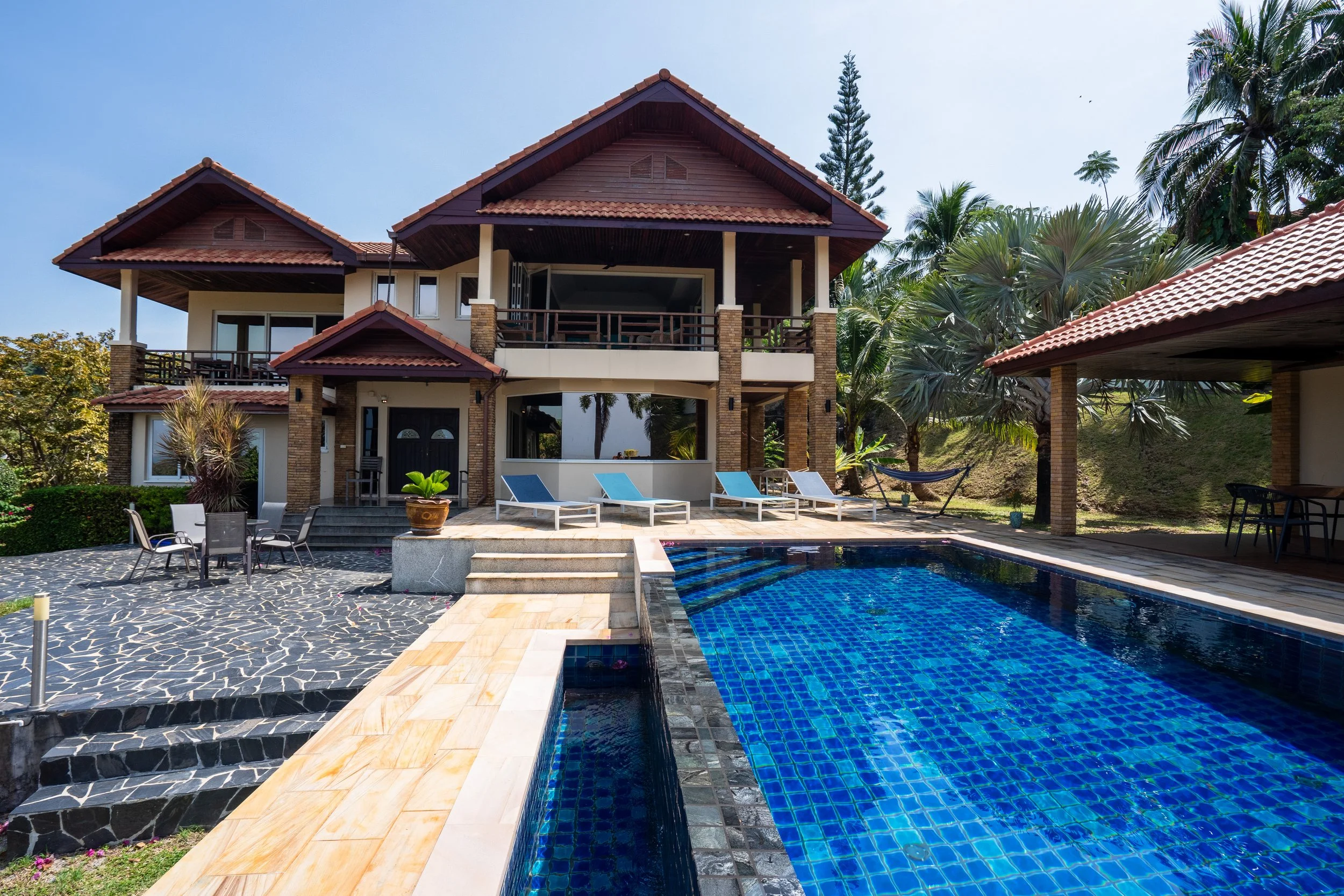 A large two-story house featuring a red tile roof and brick accents, with a swimming pool in the foreground surrounded by lounge chairs and outdoor seating, and lush greenery around.