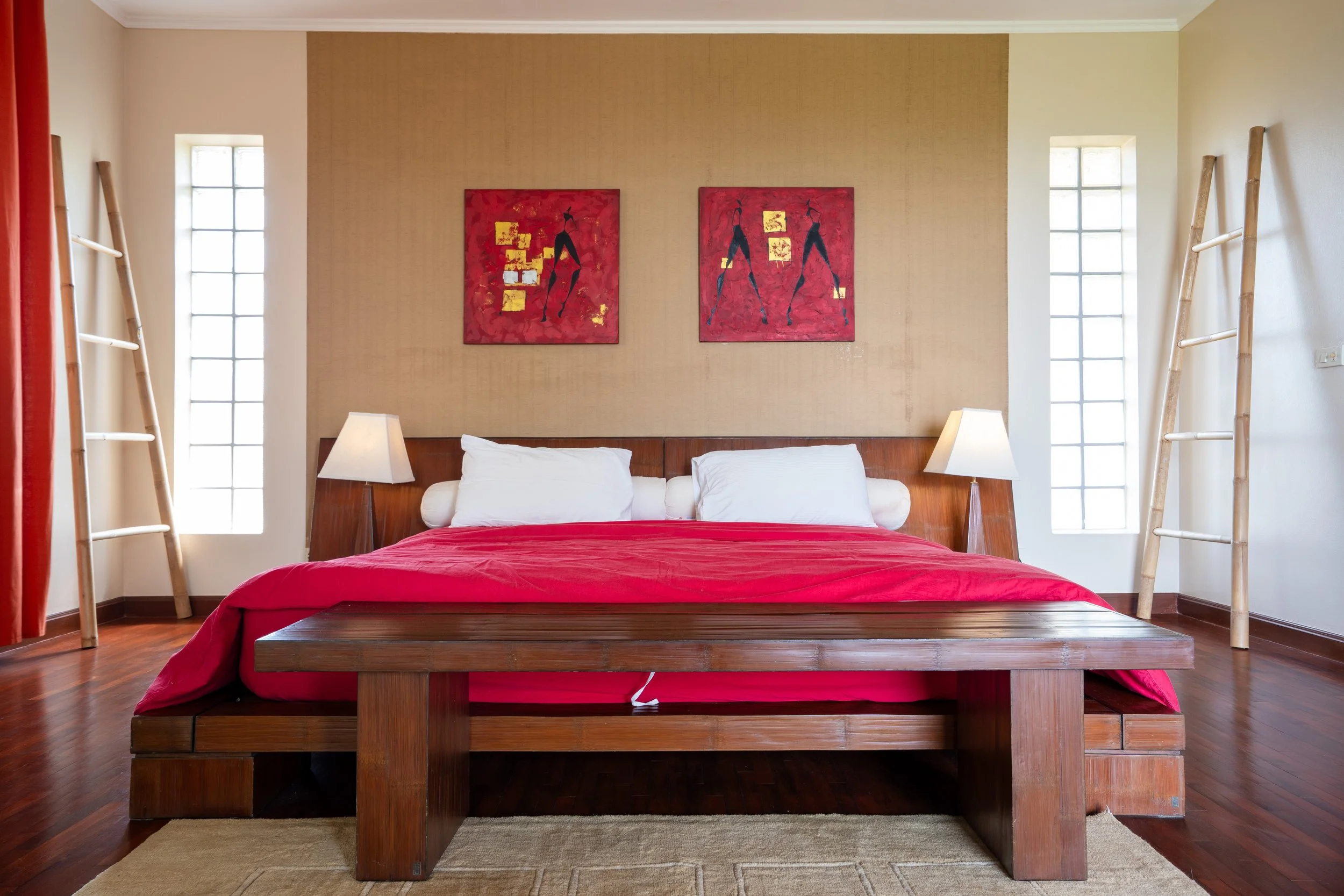 Bedroom with a wooden bed frame, white pillows, red bedding, two bedside lamps, two lattice windows, bamboo ladders on each side of the bed, and two red abstract paintings above the bed.