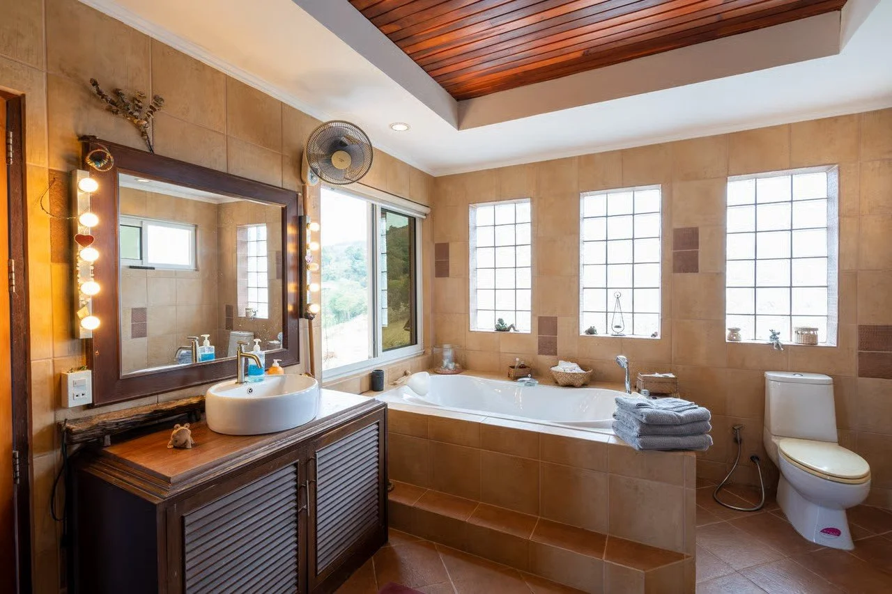 A bathroom with beige tiled walls and a wooden ceiling. It has a large corner bathtub with four windows behind it, a toilet on the right, and a wooden vanity with a round vessel sink and mirror on the left. Towels are stacked on the edge of the batht
