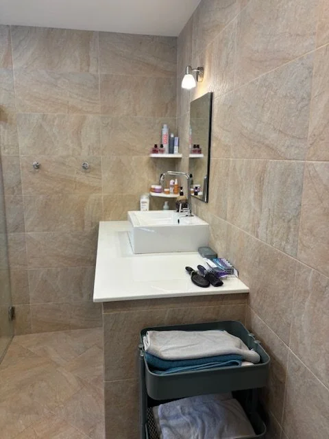 Bathroom with beige tiled walls, a white sink, a mirror, wall-mounted shelf with toiletries, and a cart with towels.