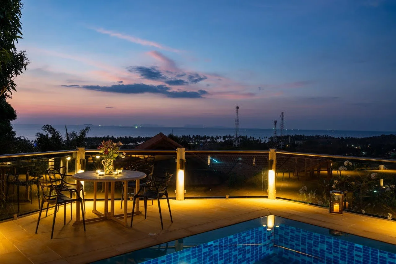 Balcony with illuminated dining table, chairs, and lanterns overlooking an ocean and evening sky with colorful clouds.