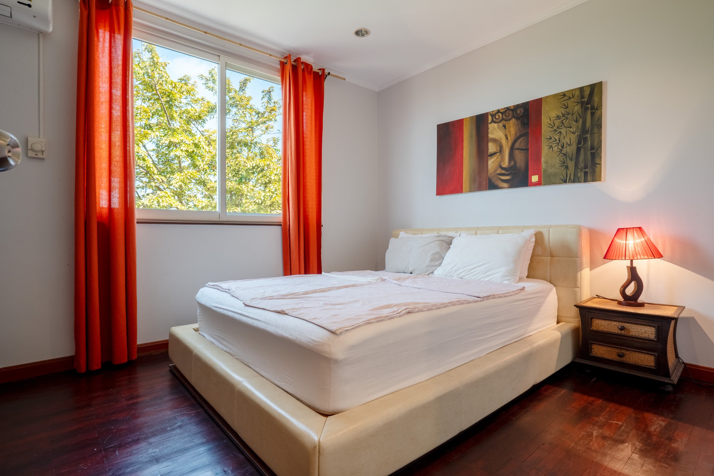 A bedroom with a large bed, white bedding, and pillows. An orange curtain on a window showing green trees outside. A bedside table with a red lamp. A colorful painting of a face and bamboo on the wall.