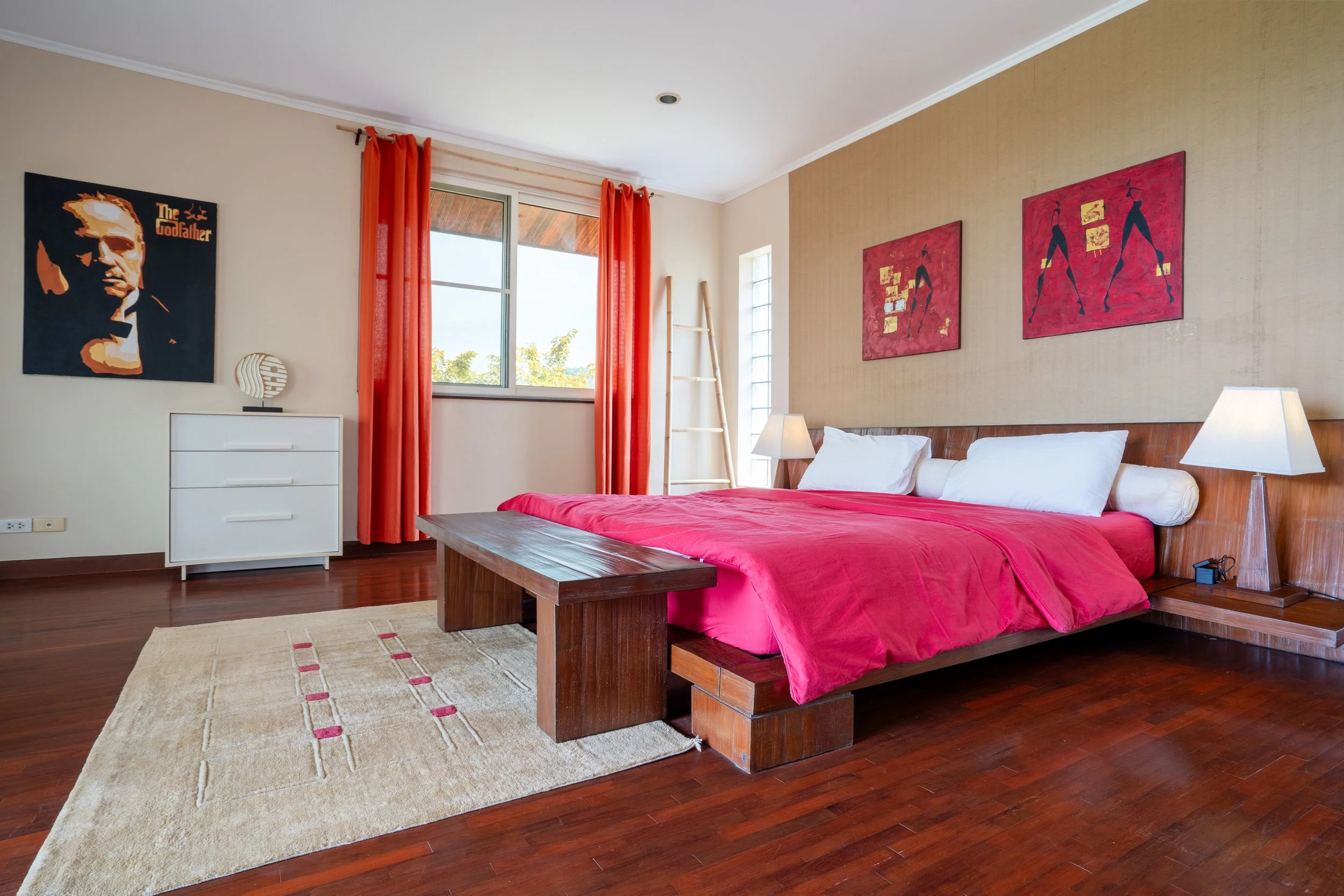 A bedroom with a large bed with a red blanket, white pillows, and a wooden headboard. There are two bedside lamps, colorful wall art, a window with orange curtains, and a white dresser. The room has wooden flooring and a beige rug.