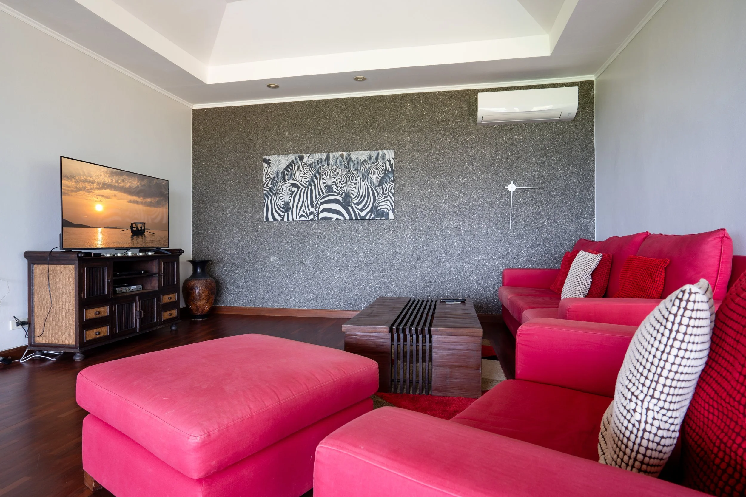 Living room with a red sofa and ottoman, a dark wooden TV stand with a flat-screen TV showing a sunset over water, a large decorative vase, a potted plant, a textured accent wall with zebra art, a modern wall clock, and an air conditioning unit.