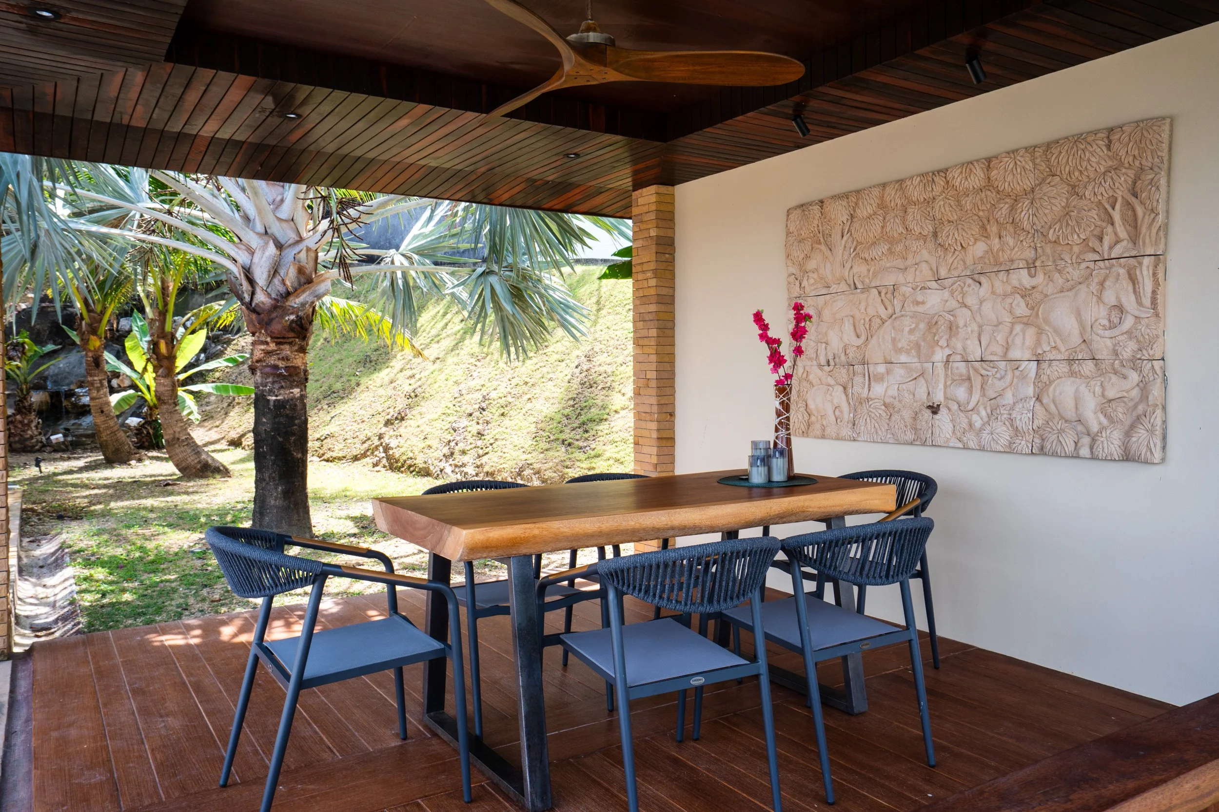 Indoor dining area with wooden table, six gray chairs, a floral centerpiece, and a carved wall art piece, next to large open window showing a tropical garden with palm trees.