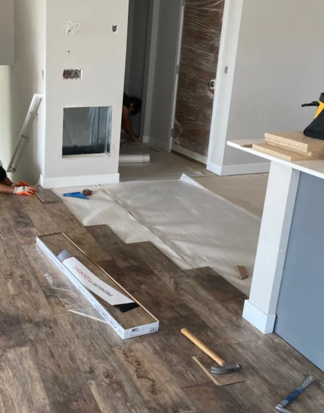 Interior of a house under renovation with workers installing wood flooring, tools scattered on the floor, and a partially covered floor area.