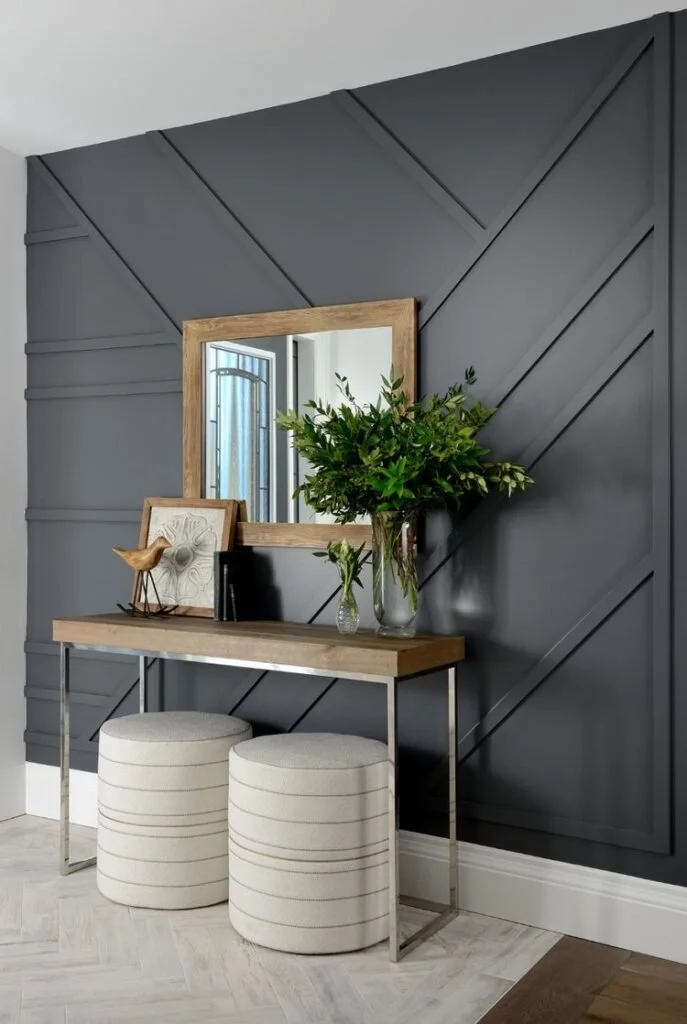 A modern entryway with a wooden console table, a large greenery arrangement in a glass vase, decorative framed artwork, a small bird figurine on a stand, two striped upholstered ottomans, a mirror on a dark gray textured wall, and light-colored wood flooring.