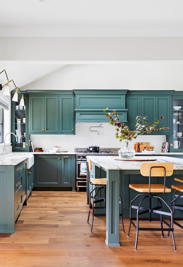 A modern kitchen with green cabinets, a white marble countertop, a central island with bar stools, a vase with branches, and a hardwood floor.