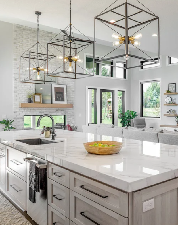 A modern kitchen with a large white marble island, black hardware, pendant light fixtures, a gray sofa, large windows, a brick fireplace, and a minimalistic decor with framed photos and plants.