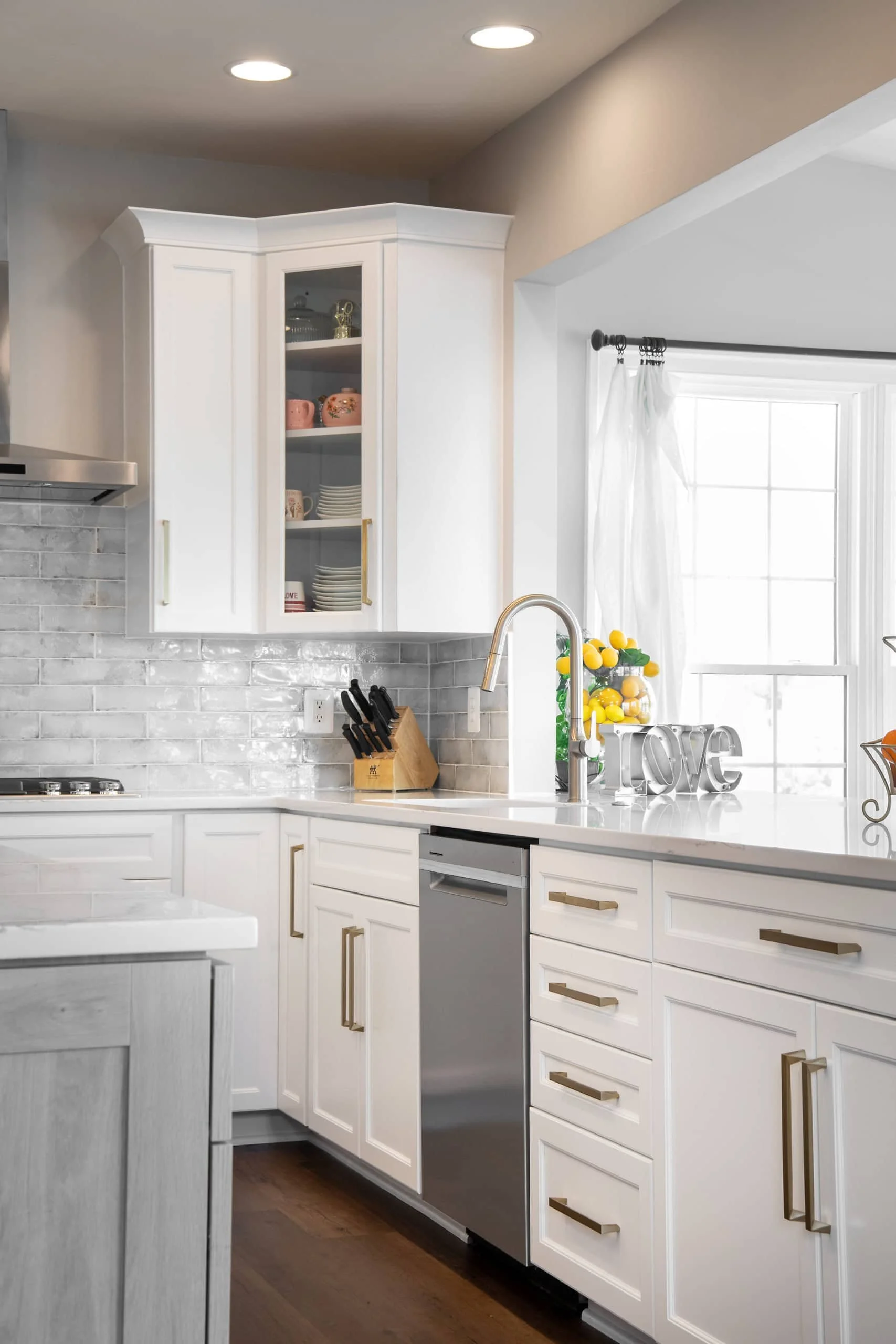 White kitchen with upper glass-front cabinets, light gray subway tile backsplash, stainless steel dishwasher, knife block, decorative lemons, and a window with white curtains.
