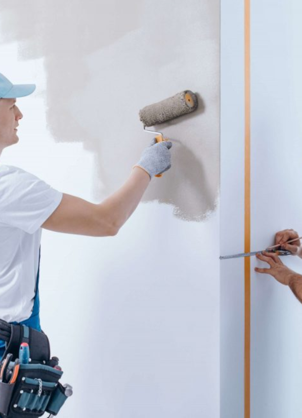 Person painting a wall with a roller brush while another person marks a line on the wall with a tape measure.