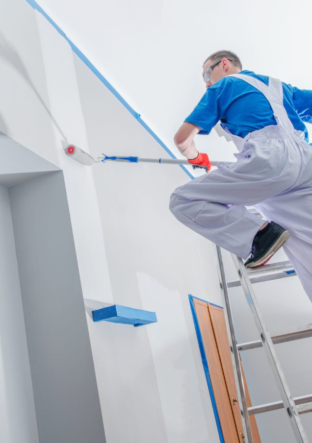 A man painting a wall white using a roller from a ladder.