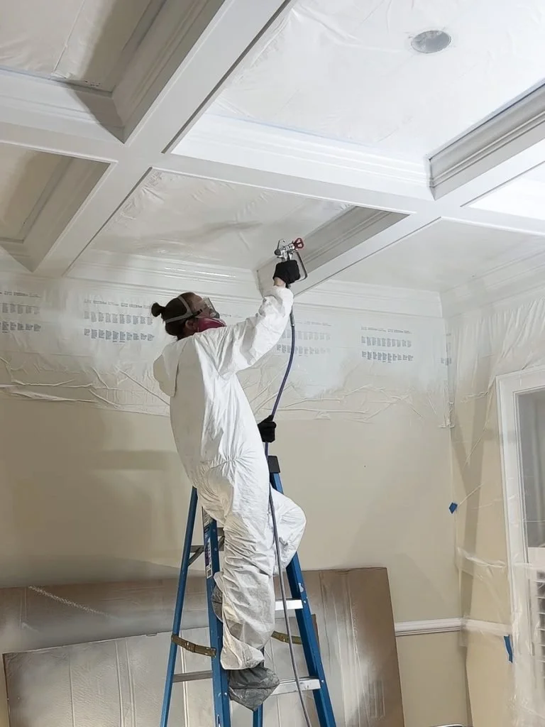 A person in a white protective suit, wearing a mask and goggles, standing on a blue ladder and spray-painting or sealing a ceiling.
