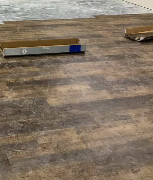 Partially unrolled flooring or wallpaper roll on a wooden floor with some cardboard packaging parts nearby.