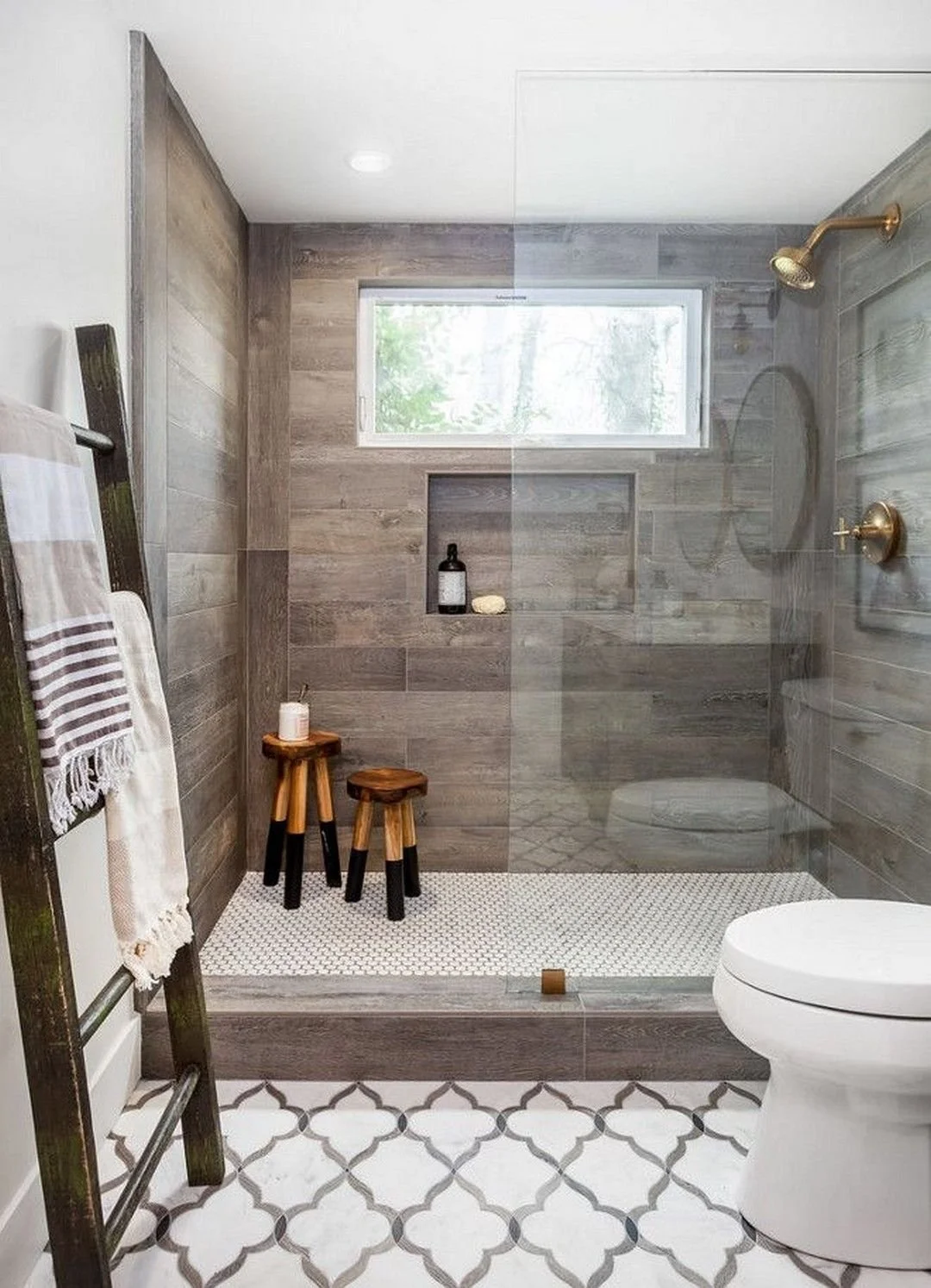 Modern bathroom with a walk-in shower featuring wood-look tiles, a small window, and brass fixtures. The shower has a built-in niche with toiletries, and the bathroom floor has a white and gray patterned tile. There is a white toilet and a wooden towel rack with towels.