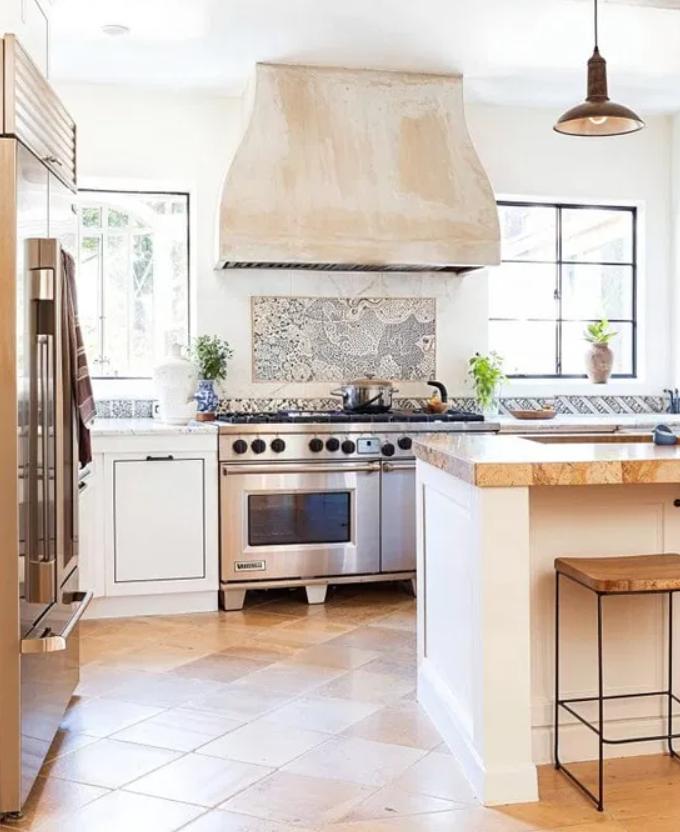 Modern kitchen with stainless steel stove and oven, white cabinetry, a large range hood, windows with plants, and a tiled backsplash.