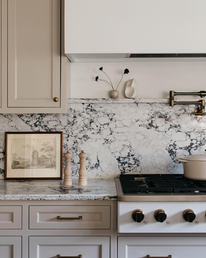 Kitchen with white cabinetry, marble countertop and backsplash, framed artwork, salt and pepper shakers, and a stove with a pot on top.