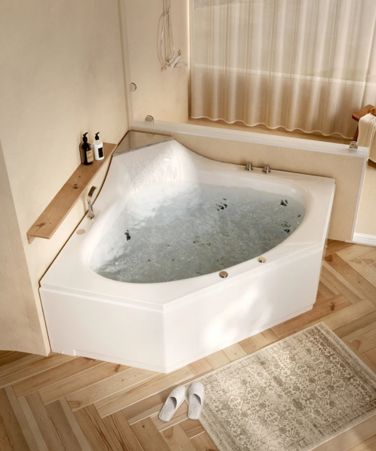 A white soaking bathtub filled with water, placed in a cozy corner with beige curtains and wooden accents. Bath products sit on a wooden shelf beside the tub. White slippers rest on a beige rug on a hardwood floor.
