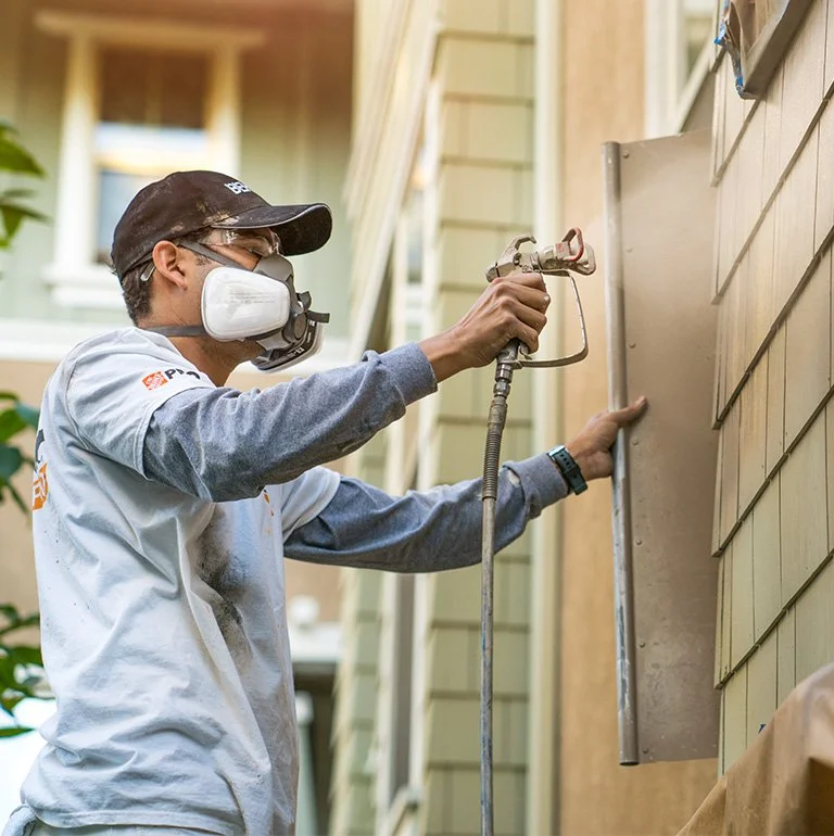 A person in safety gear spray painting the exterior wall of a house.