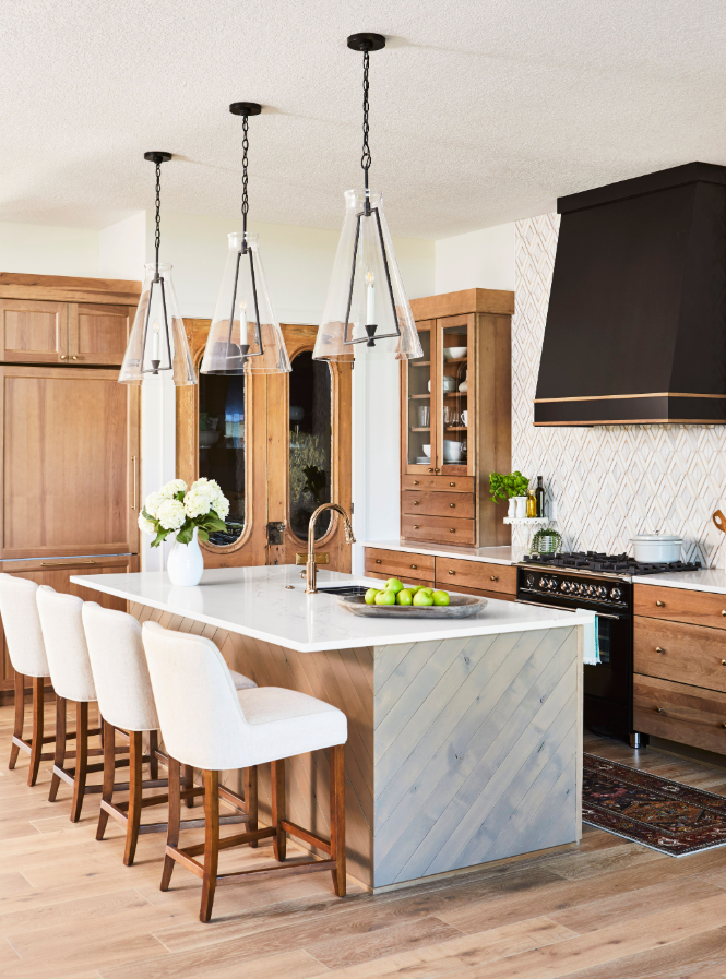 Modern kitchen with wood cabinets, a white island with a marble countertop, four white upholstered chairs, pendant lighting, and a black range hood.