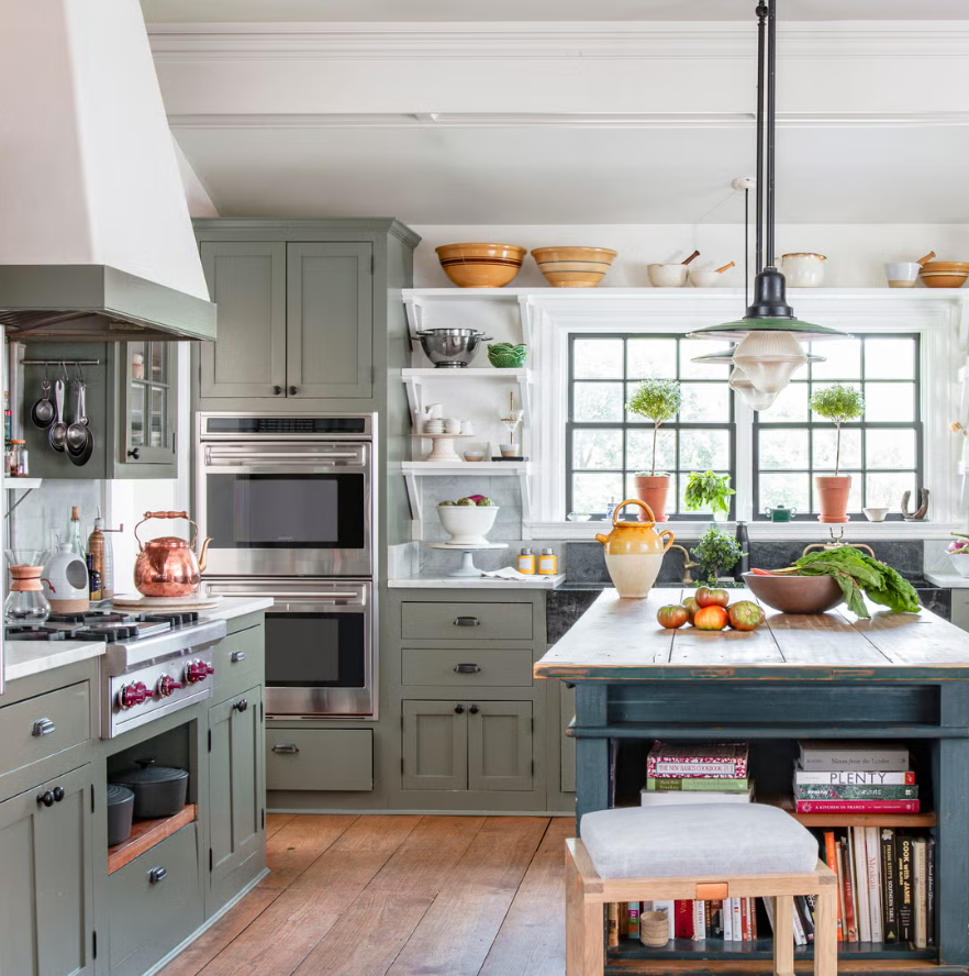 A cozy, well-organized kitchen with a large island, green cabinetry, open shelves, a window with potted plants, and various kitchen accessories.