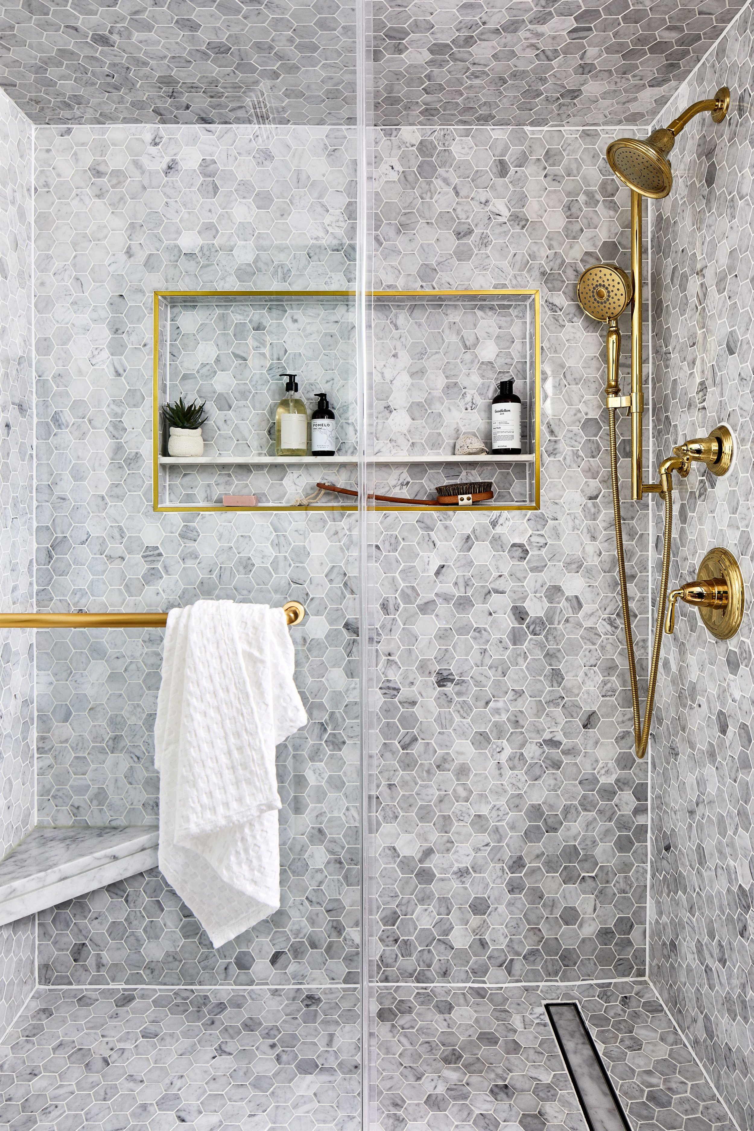 A modern walk-in shower with hexagonal gray tiles, two shower heads, a built-in shelf with toiletries, a towel rack with a white towel, and a floor drain.