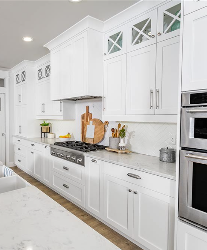 Modern white kitchen with marble countertops and upper cabinets with glass doors, featuring a stovetop, cutting boards, and kitchen utensils.