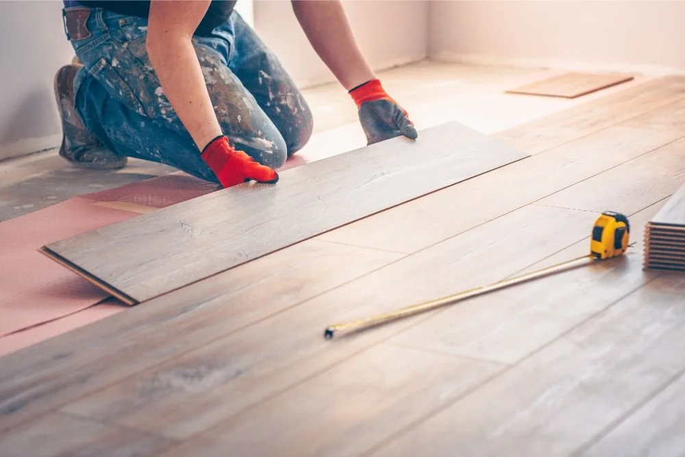 Worker installing wood flooring, kneeling on the floor with a tape measure and flooring planks.