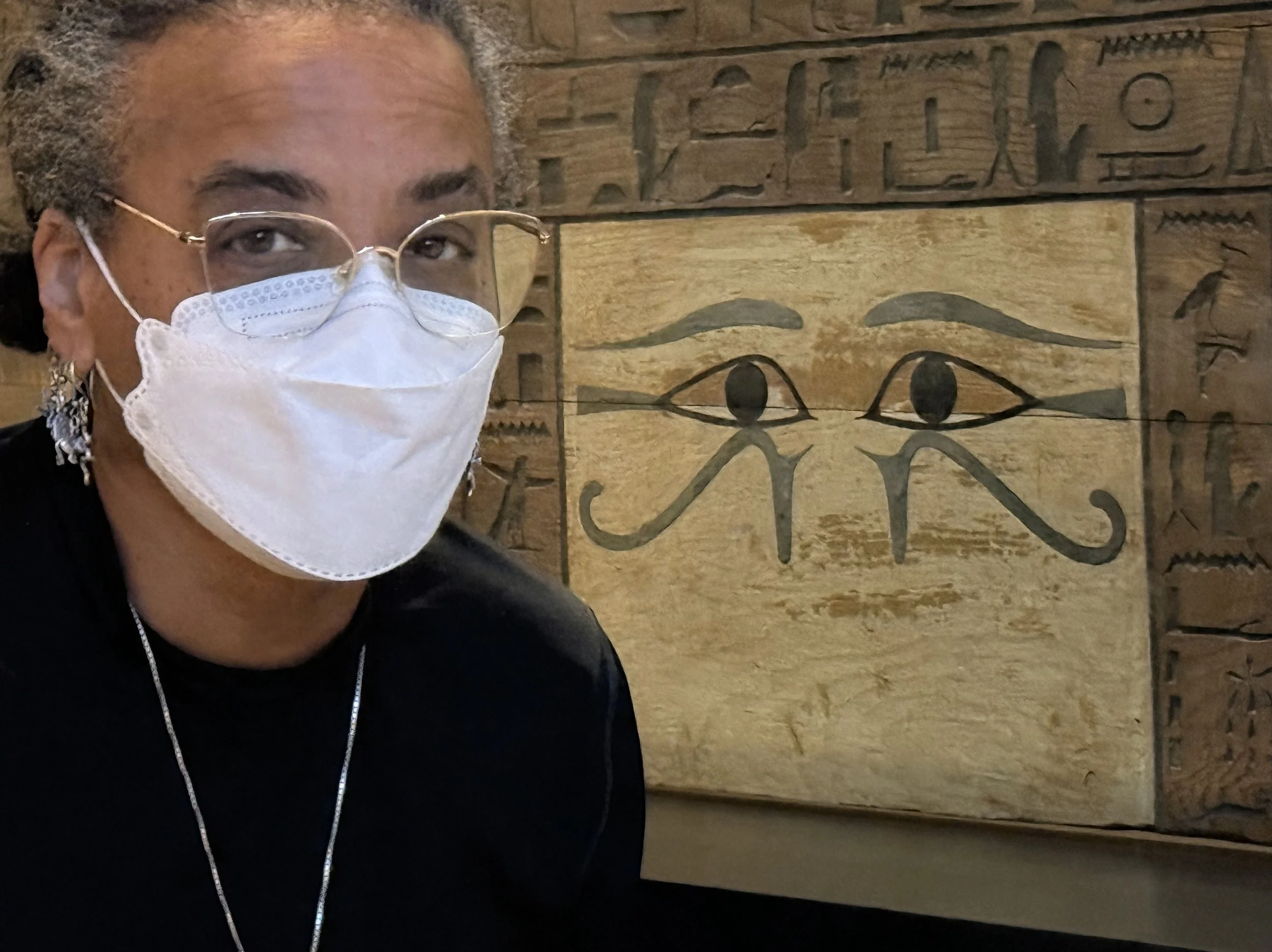 Ali Montgomery in N95 mask standing before a wooden coffin panel carved with hieroglyphs and painted with two prominent Udjat eyes, at the Kunsthistorisches Museum in Vienna.