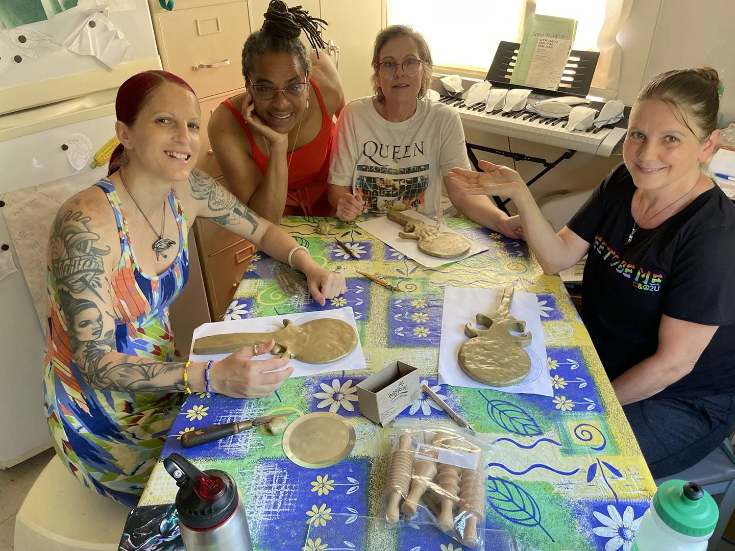Four women seated around a table with partially completed gold‑colored clay mirrors and sculpting tools.